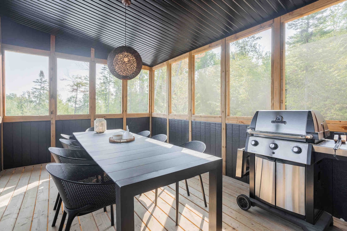 A screened-in balcony features a large dining table surrounded by black wicker chairs. A stainless steel barbecue is positioned nearby. Natural light filters through the numerous large windows, highlighting the wooden flooring and the cozy atmosphere.