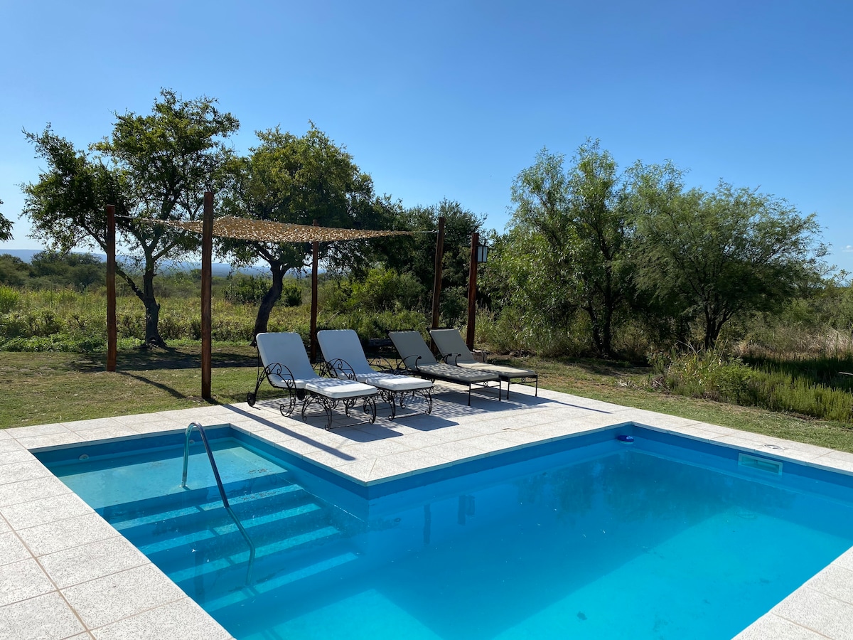 A well-appointed outdoor pool area features a sparkling blue swimming pool surrounded by a spacious deck. Comfortable lounge chairs are arranged nearby, while natural greenery provides a serene backdrop. A shaded pergola offers a place for relaxation under the sun.