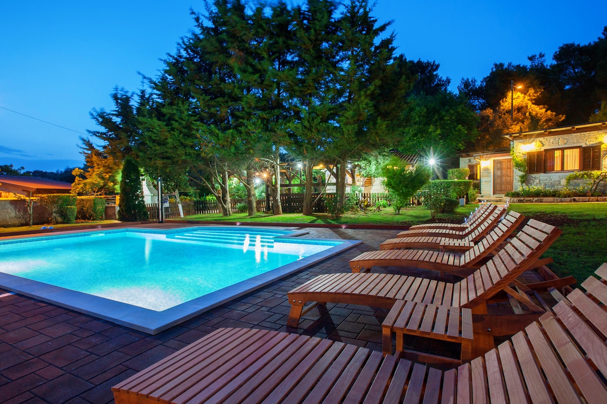 A serene outdoor space is captured at dusk, featuring a rectangular swimming pool illuminated in bright blue. Sun loungers are arranged nearby, surrounded by lush greenery and trees. The structure in the background displays warm lighting, offering a welcoming ambiance.