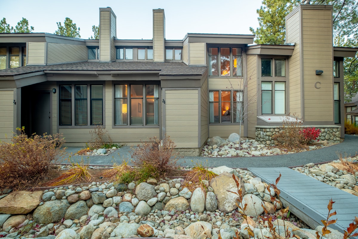 The exterior of a modern townhome is displayed, featuring a combination of wood and stone accents. Large windows provide ample light, while well-maintained landscaping and a rock pathway lead to the entrance, enhancing the natural setting.