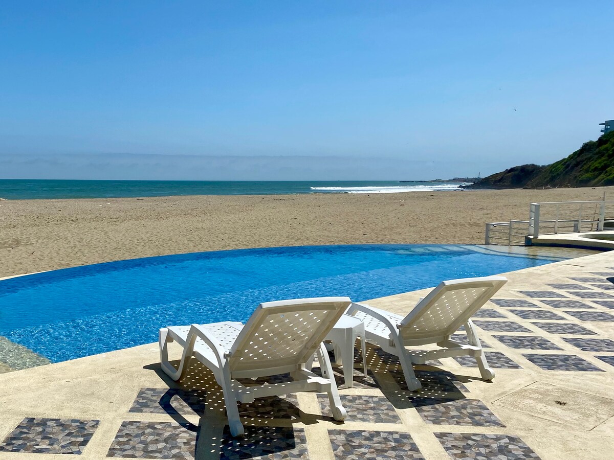 A serene infinity pool extends towards the coastline, framed by inviting lounge chairs. The golden sandy beach meets the ocean waves, while the clear blue sky enhances the tranquil setting. Nearby, a gentle slope leads up to green hills, creating a natural backdrop.