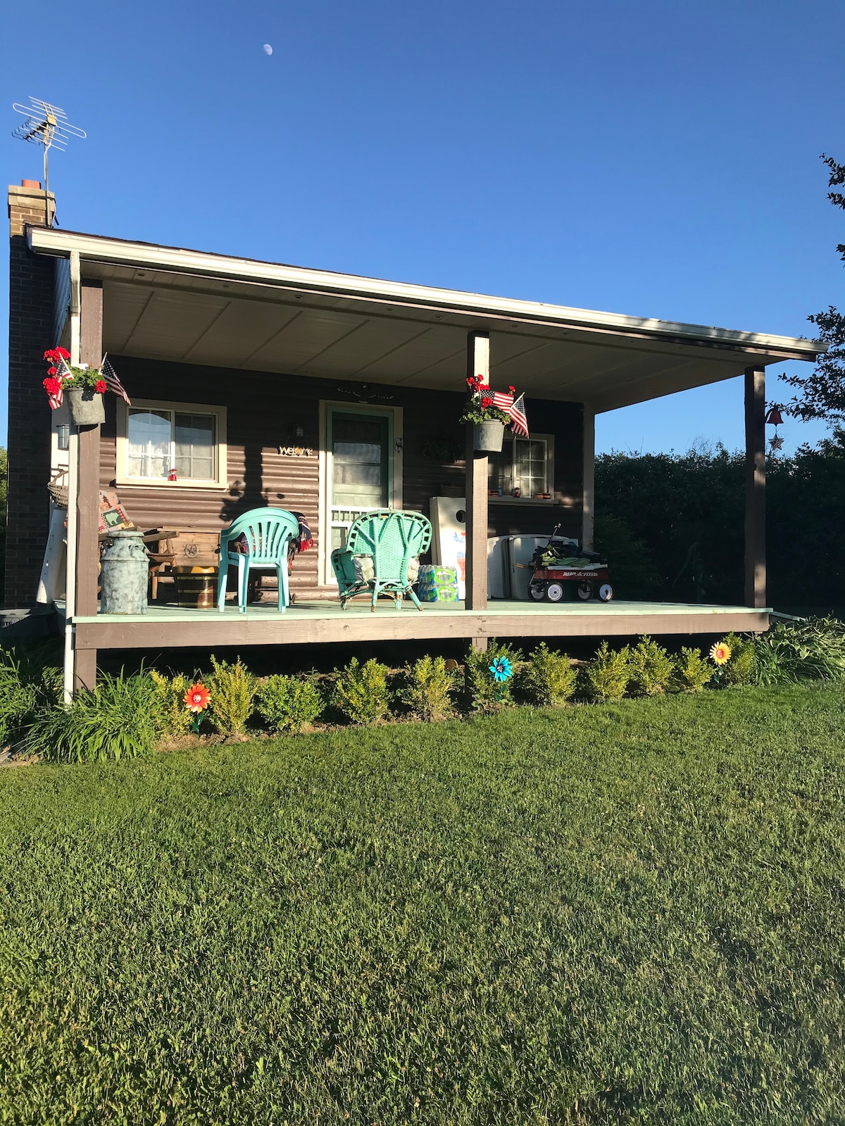 The front porch features two light blue chairs and decorative potted plants. A green rug and small cart add charm, while colorful flowers line the edge of the grassy area. The warm wooden cabin structure is complemented by the clear blue sky.