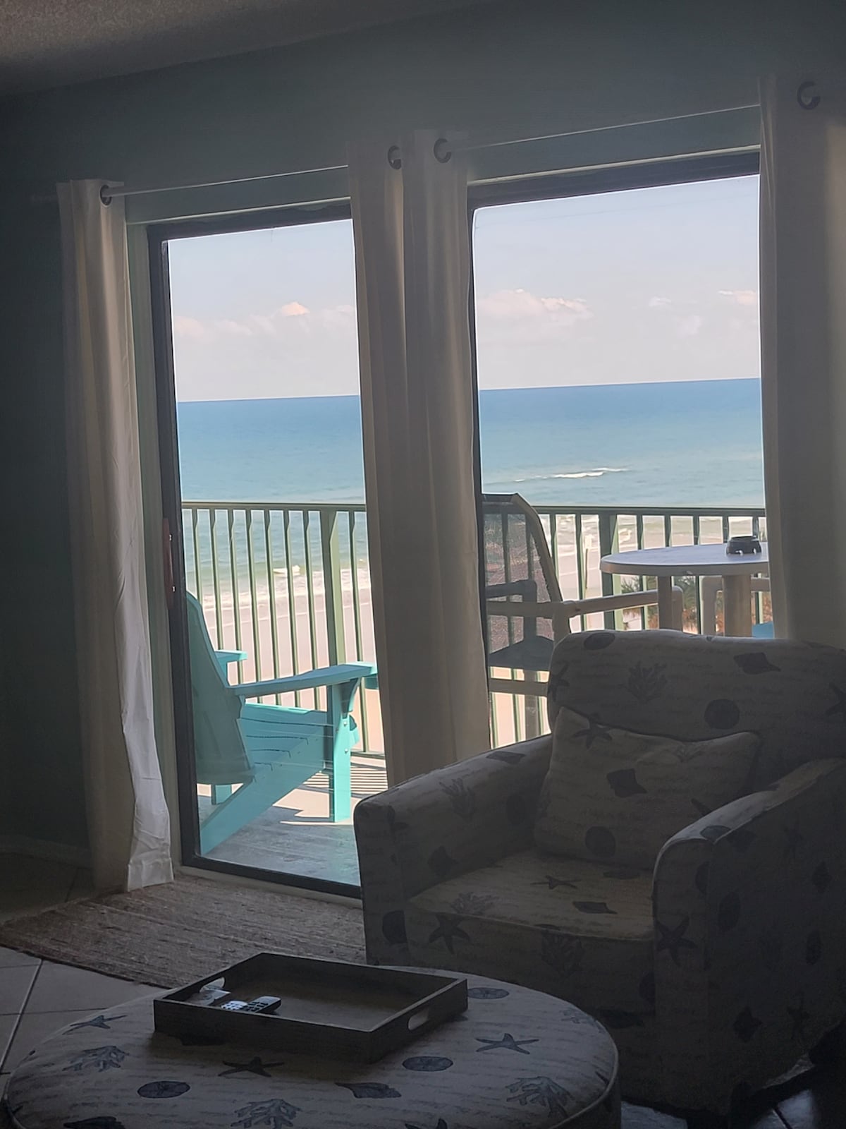 A sliding glass door frames a serene ocean view, with gentle waves visible in the distance. An inviting chair in the foreground complements the coastal decor, while light curtains softly filter natural sunlight, enhancing the calming atmosphere of the space.