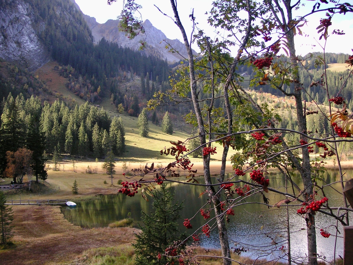 A serene landscape is portrayed, featuring a tranquil lake surrounded by lush vegetation and autumn foliage. The mountains rise in the background, and the reflections on the water create a peaceful scene. A few trees are adorned with red berries, enhancing the natural beauty.