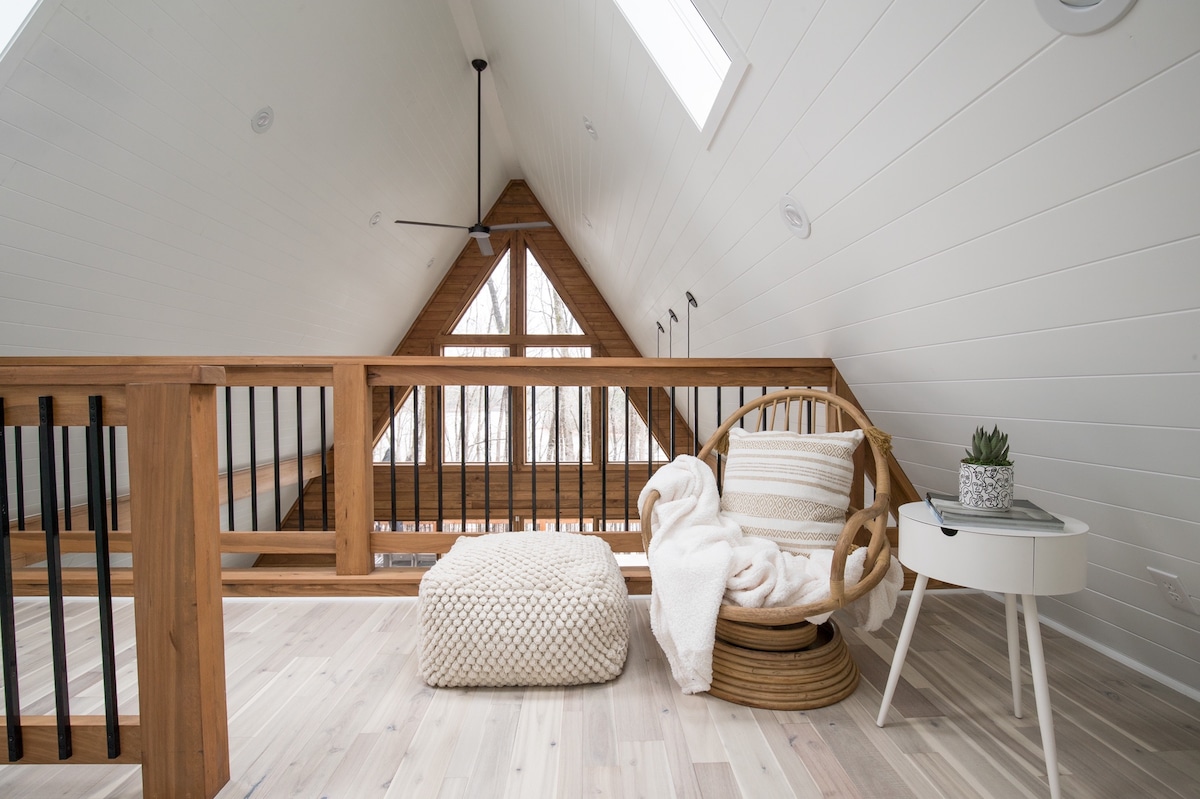 A cozy loft area features wooden railings and sloped ceilings adorned with skylights. A circular chair with soft cushions is accompanied by a pouf and a small table. Natural light enhances the serene atmosphere, creating a perfect reading nook.