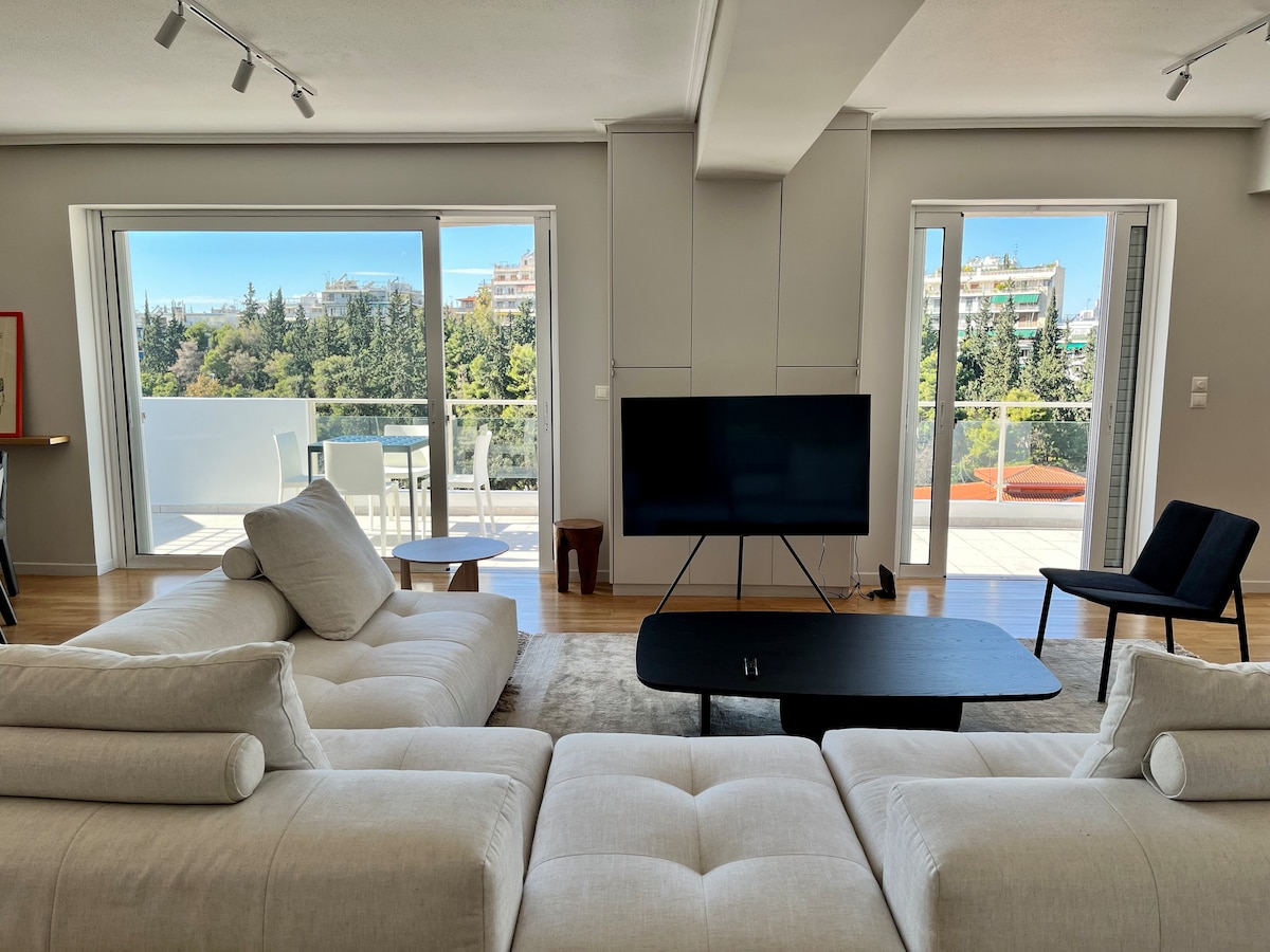 The living room features a spacious sofa facing a coffee table and a large smart TV. Expansive glass doors lead to a balcony with city views. Natural light fills the room, creating an airy atmosphere, complemented by minimalist decor.