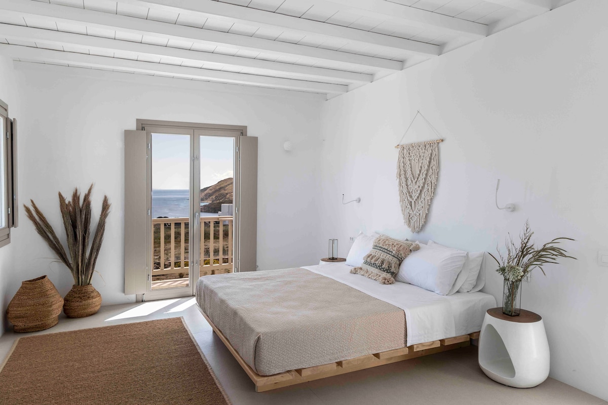 A spacious bedroom features a large bed with neutral bedding, complemented by natural decor elements. Two doors open to a balcony, offering an unobstructed sea view. Minimalist furnishings, including a round bedside table and decorative baskets, enhance the serene ambiance of the room.