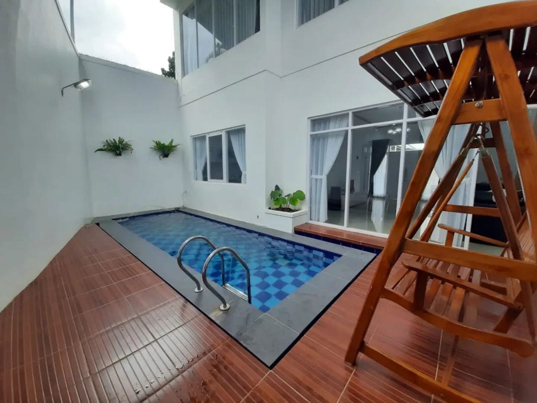 A private pool area is set against a modern white wall, featuring a ladder for easy access. Surrounding foliage adds a touch of greenery, while large windows provide natural light. A wooden swing is positioned nearby, enhancing the outdoor space.