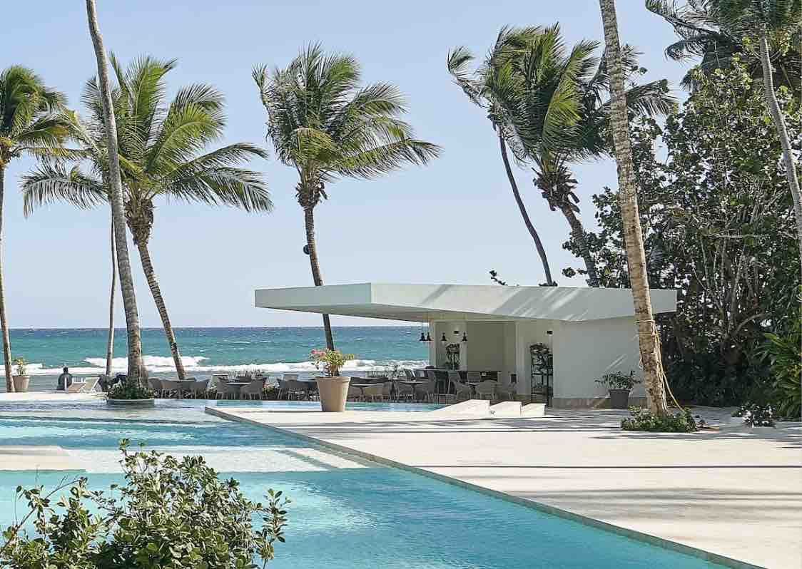 A modern structure is seen near a sparkling pool, surrounded by lush palm trees and facing the ocean. Clear skies reflect off the water, creating a serene atmosphere. Outdoor seating is available near the pool, offering a panoramic view of the coastline.