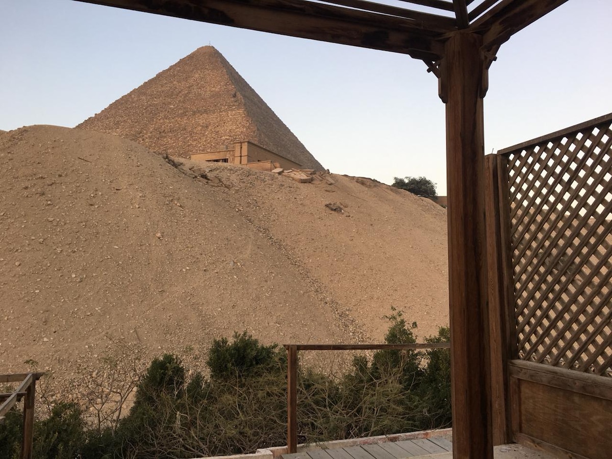 A large pyramid is prominently displayed in the foreground, showcasing its distinct, sloping sides and stone structure. The scene is framed by wooden beams and a lattice, with greenery visible at the base, enhancing the connection to the outdoor environment.