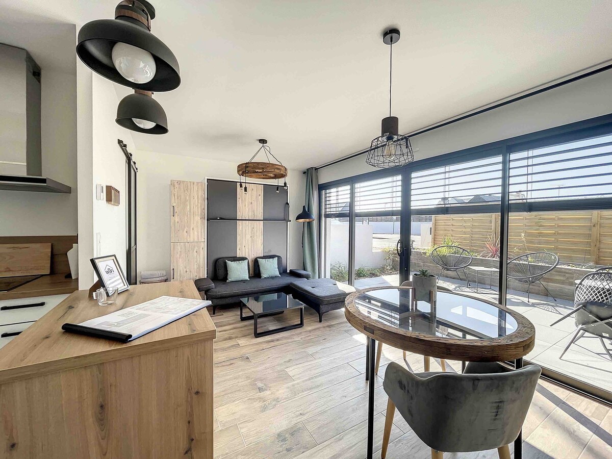 An open living space is presented with large glass doors offering outdoor access. A dark sofa and a round dining table with chairs create a comfortable area. The flooring is light wood, and stylish pendant lights hang from the ceiling, enhancing the modern decor.