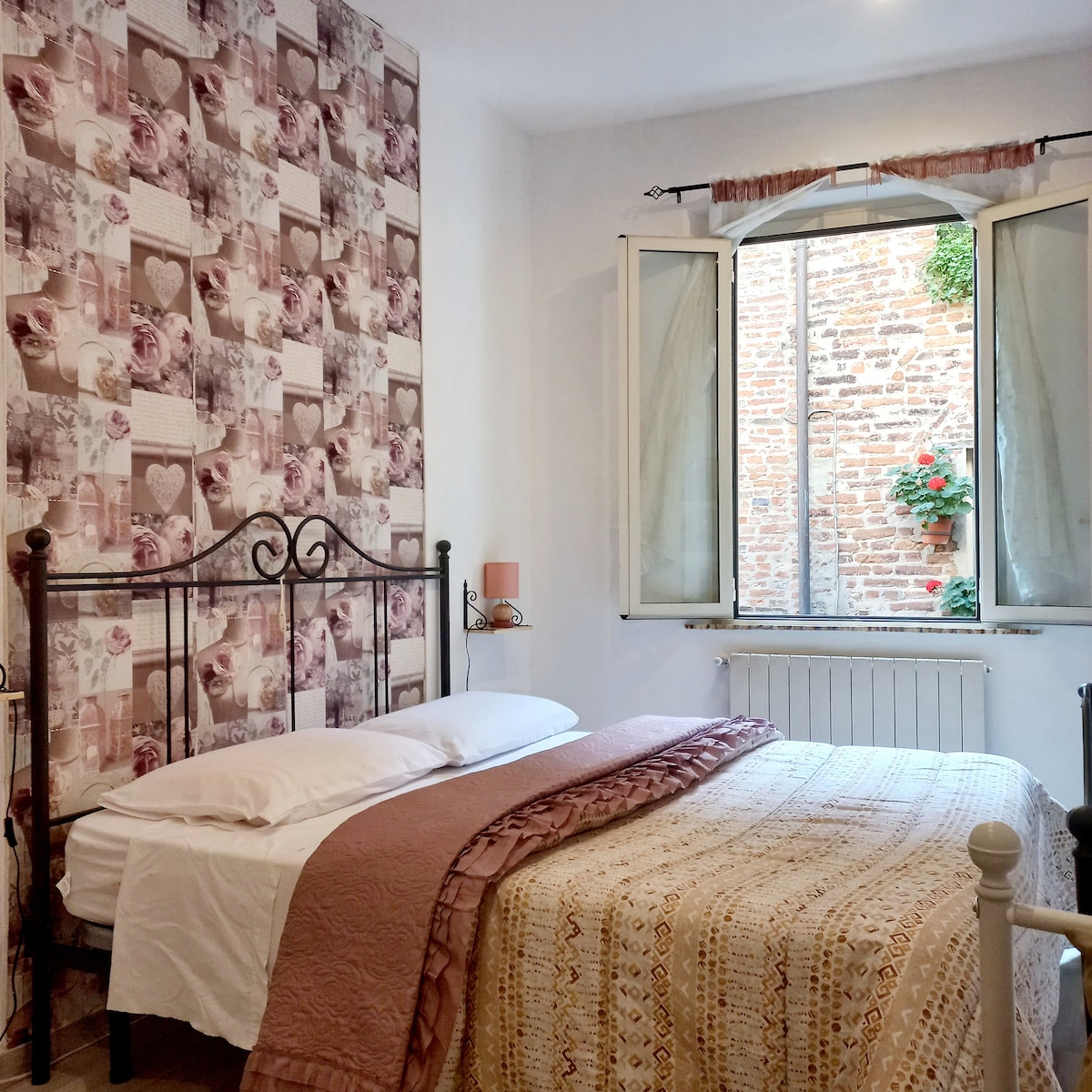A charming bedroom features a wrought iron bed with a lightweight blanket draped over it. Natural light enters through a large window with open shutters, revealing a view of brick walls adorned with plants. Floral wallpaper adds a subtle touch to the room's decor.