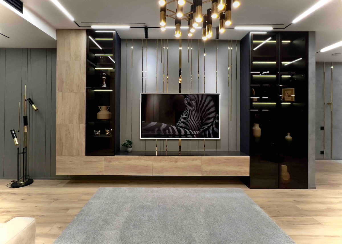 A modern living area is depicted, featuring a sleek entertainment unit with a large flat-screen TV mounted on the wall. Gold accents are highlighted in the cabinetry and light fixtures. A soft gray rug covers the floor, contributing to a contemporary aesthetic.