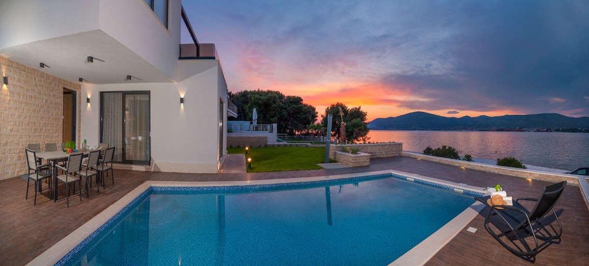 An outdoor area features a blue swimming pool bordered by a wooden deck, with lounge chairs positioned nearby. A dining set with several chairs is seen to the left, while surrounding gardens and the sunset sky reflect vibrant colors over the sea.