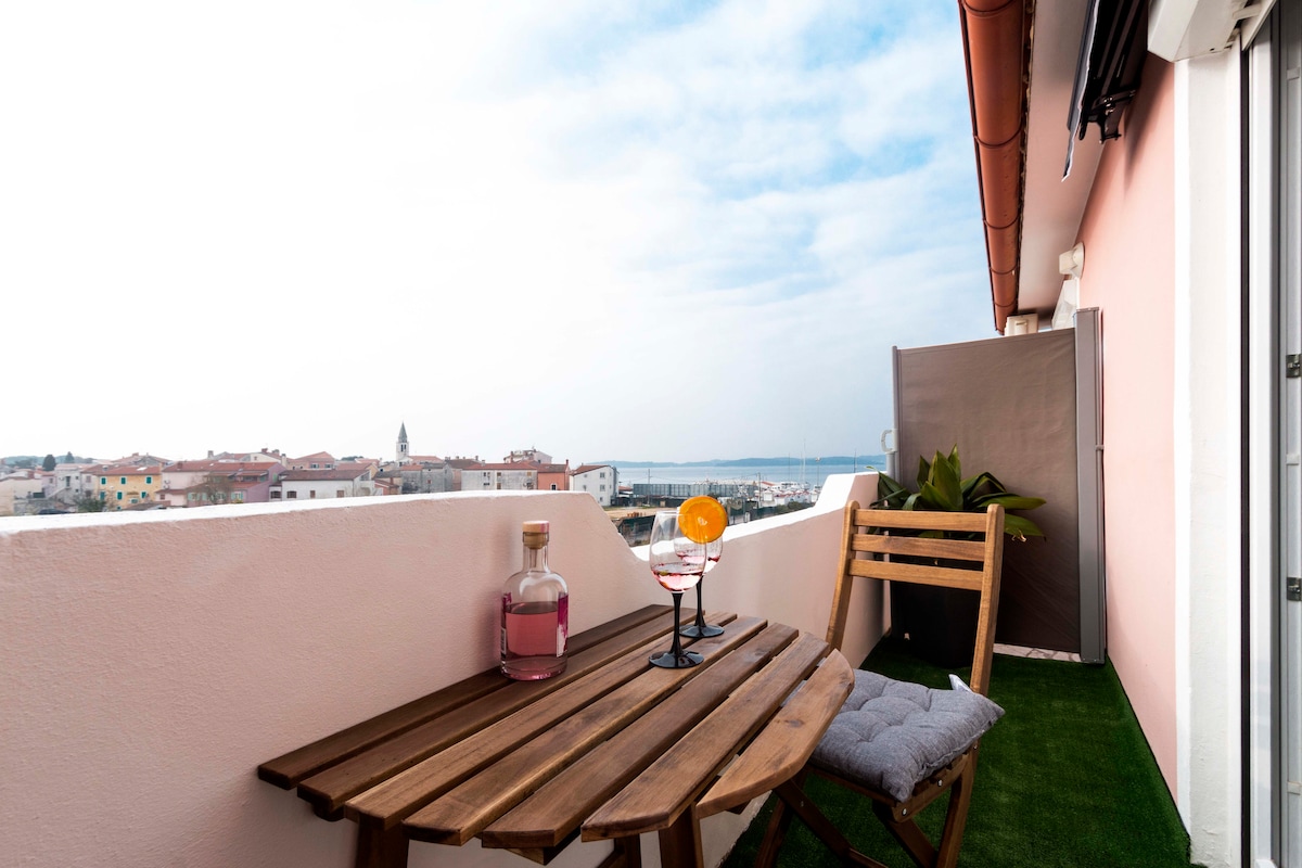 A small balcony features a wooden table with two glasses and a carafe, surrounded by greenery. A folding chair provides seating. The view includes the town and hints of the sea under a cloudy sky, creating a serene outdoor space.