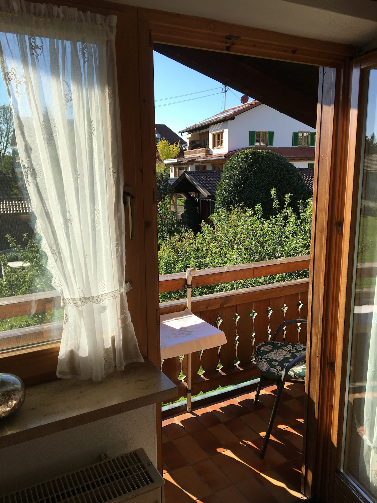 A view from the apartment's window reveals a sunny balcony with a small table set against a leafy backdrop. Natural light filters through a sheer white curtain, highlighting the wooden frame and tiled floor. A nearby house can be seen through the greenery.