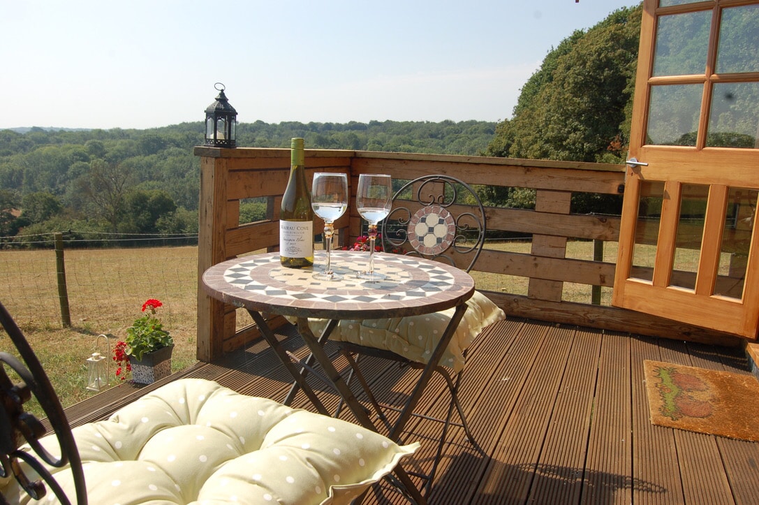 An outdoor terrace features a small circular table adorned with a patterned top, accompanied by two glasses and a bottle of wine. Comfortable cushions are placed on the metal chairs, offering a view of the surrounding countryside and trees beyond the railing.