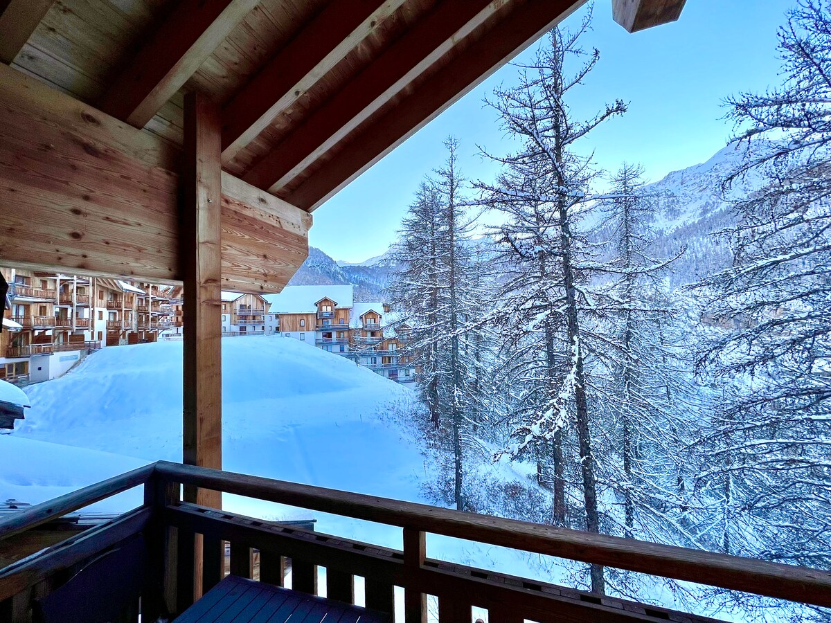 A winter landscape is captured from the balcony, showcasing snow-covered hills and trees. The surrounding chalets are partially visible, blending with the serene white scenery. The wooden balcony railings add a rustic touch, enhancing the view of the tranquil environment.