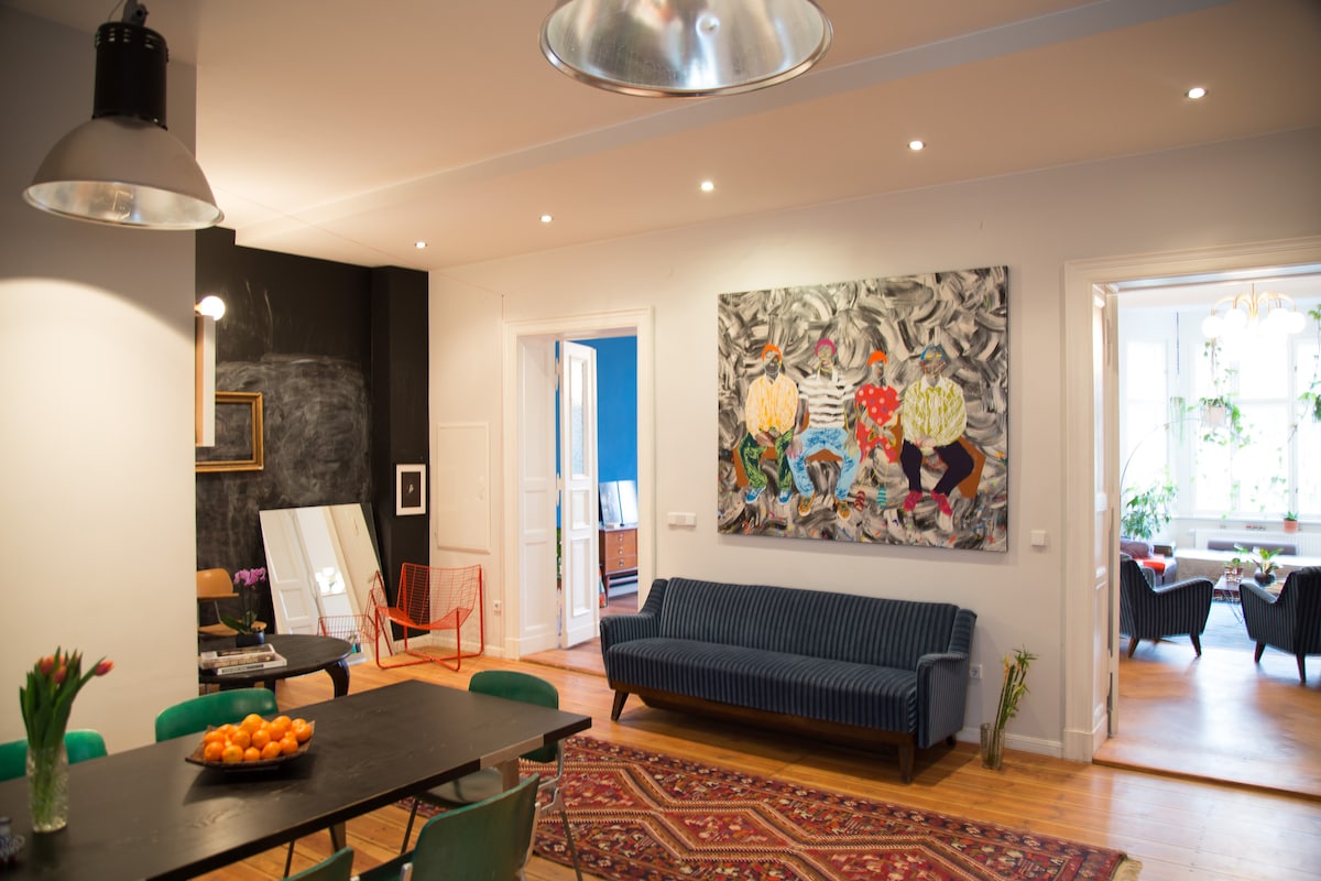 An open living area features a long dining table with green chairs and a bowl of oranges. A navy blue sofa is positioned near a large colorful artwork mounted on the wall, while illuminated recessed lighting adds brightness. The space includes an accent wall and various seating arrangements.