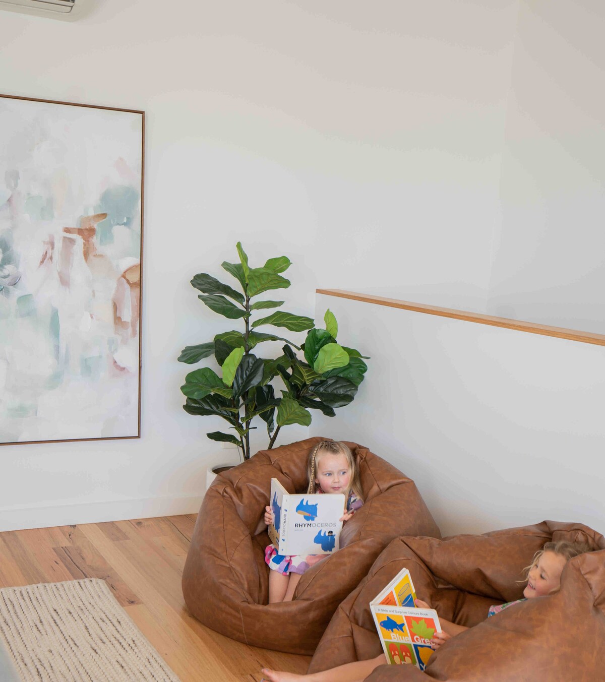 A cozy nook is depicted with two large bean bag chairs positioned near a leafy green plant. A vibrant artwork hangs on the wall, complementing the light wood flooring. A child is seen occupying one of the bean bags, focused on reading a book.