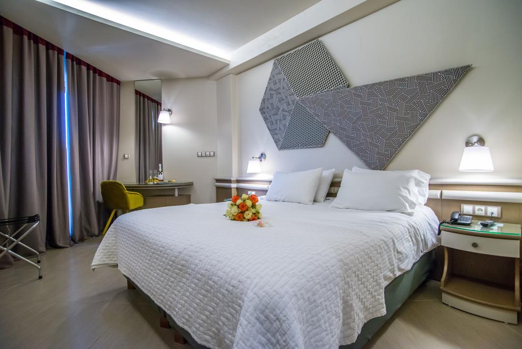 A spacious room is furnished with a large bed topped with a decorative arrangement of flowers. A desk with a chair sits nearby, facing a wall featuring patterned artwork. Soft lighting is provided by wall-mounted lamps beside the bed, with sheer curtains allowing natural light to filter in.