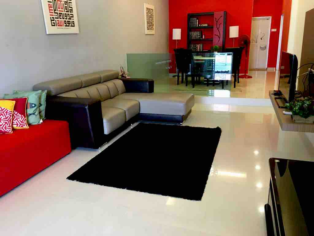 A modern lounge area features a contemporary grey and black sectional sofa on a plush black rug. A glass coffee table sits on the polished floor, with a bright red accent wall providing contrast. Behind, a dining area is visible, complemented by minimalist decor.