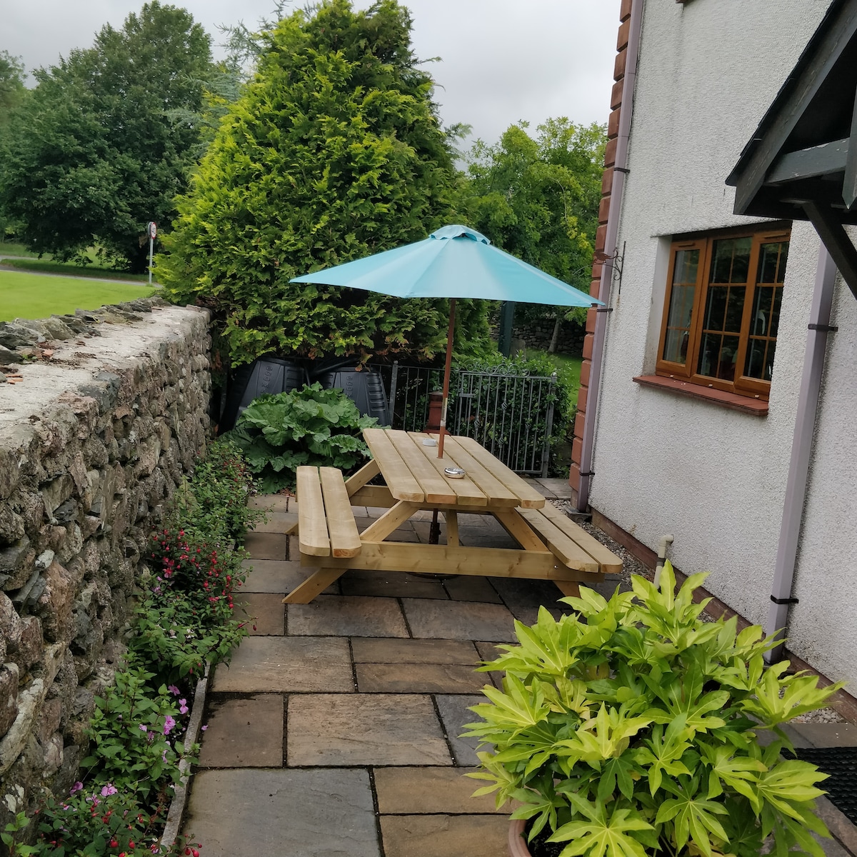 An outdoor seating area is featured, showcasing a wooden picnic table beneath a light blue umbrella. Surrounding greenery is evident, including well-maintained shrubs and flowers along stone pathways, with a stone wall in the background adding texture to the scene.