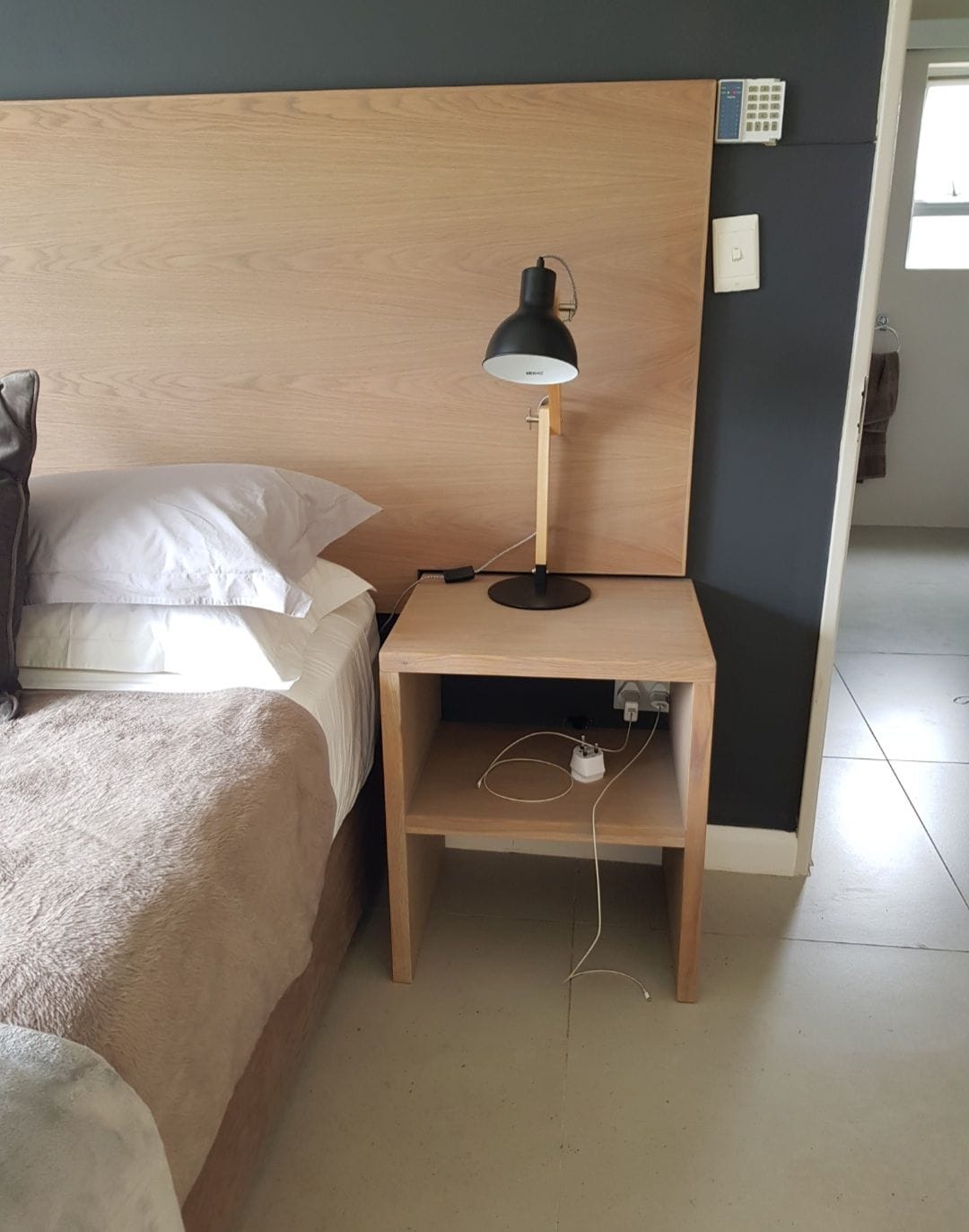 A neatly arranged sleeping area features a spacious bed with light-colored bedding and a warmly textured throw. A minimalist nightstand is positioned alongside the bed, equipped with a sleek lamp and accessible charging outlets. A doorway leading to a bathroom is visible in the background.
