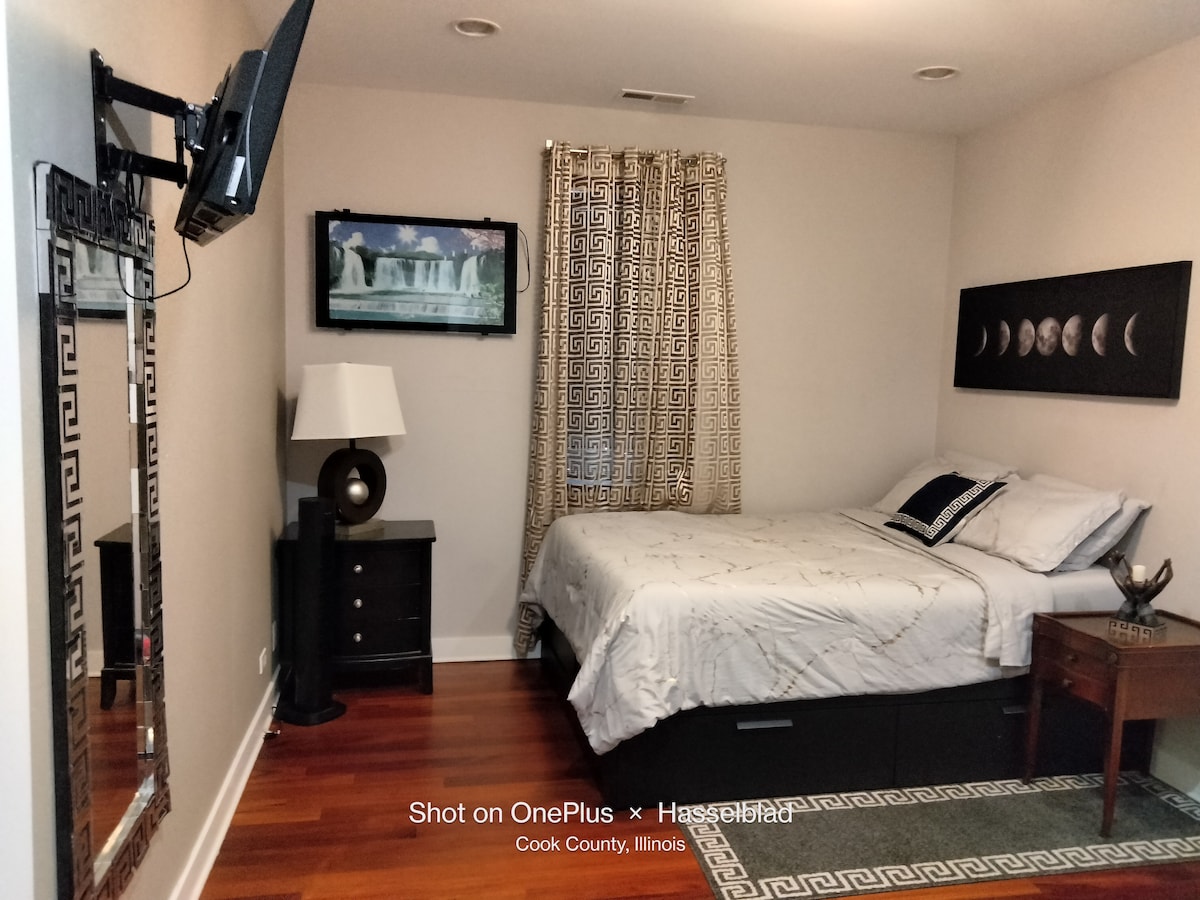 An elegant bedroom features a spacious bed with decorative bedding. A mounted TV is positioned on the wall opposite the bed. A stylish mirror and bedside table with a lamp are included, complemented by window treatments that introduce a subtle pattern. Warm hardwood flooring completes the space.