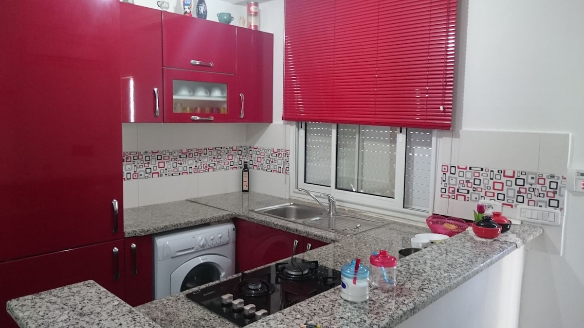 A modern kitchen features sleek red cabinetry and a granite countertop. A stainless steel sink is positioned near a window adorned with red blinds. A washing machine is tucked into a corner, and colorful decorative items are arranged along the countertop.