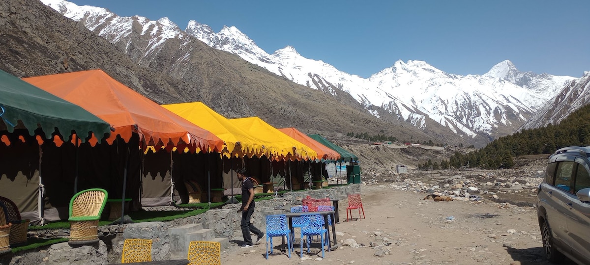 Colorful tents in vibrant orange and yellow are arranged along a mountainous landscape, framed by snow-capped peaks. Outdoor tables in blue and pink are set nearby, providing a space for relaxation amidst nature. The scene captures the serene beauty of the surrounding environment.