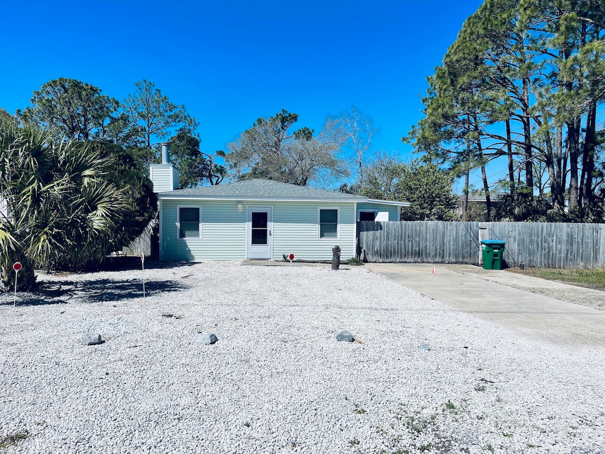 The exterior of the house is shown, featuring a spacious gravel driveway and a small landscaped area with palm trees. A fenced yard is visible, providing privacy. The house displays a light-colored facade with multiple windows, set against a clear blue sky and natural greenery.