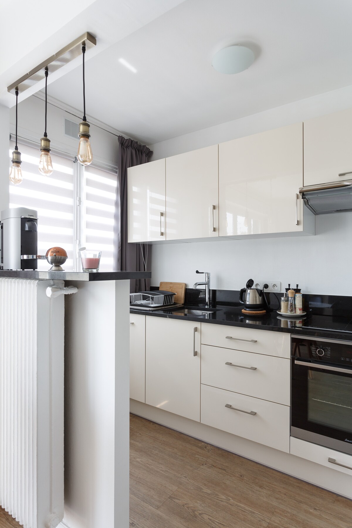 The modern kitchen features sleek white cabinetry and a dark countertop. Stainless steel appliances, including a coffee maker and kettle, are positioned for convenience. Natural light enters through a window dressed with light-filtering blinds, enhancing the functional design of the space.