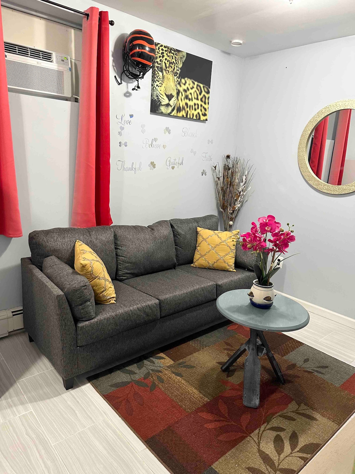 A cozy lounge area features a gray sofa with decorative yellow pillows, accompanied by a round table. A small floral arrangement sits atop the table. The walls are decorated with a leopard print motif and inspirational quotes, complemented by vibrant red curtains.