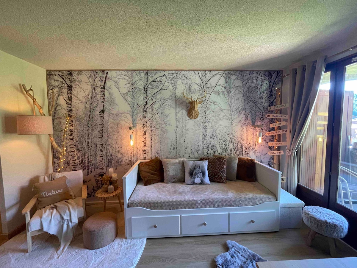 A cozy living area is furnished with a spacious daybed featuring an integrated storage drawer. The wall is adorned with a serene forest mural, and soft lighting is provided by wall sconces. A comfortable chair and decorative elements enhance the welcoming ambiance.