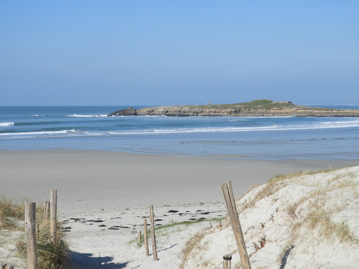A sandy beach stretches towards the horizon, meeting gentle waves under a clear blue sky. The shoreline is framed by grassy dunes, with wooden posts marking the path to the water. A distant rocky outcrop is visible, adding interest to the coastal landscape.