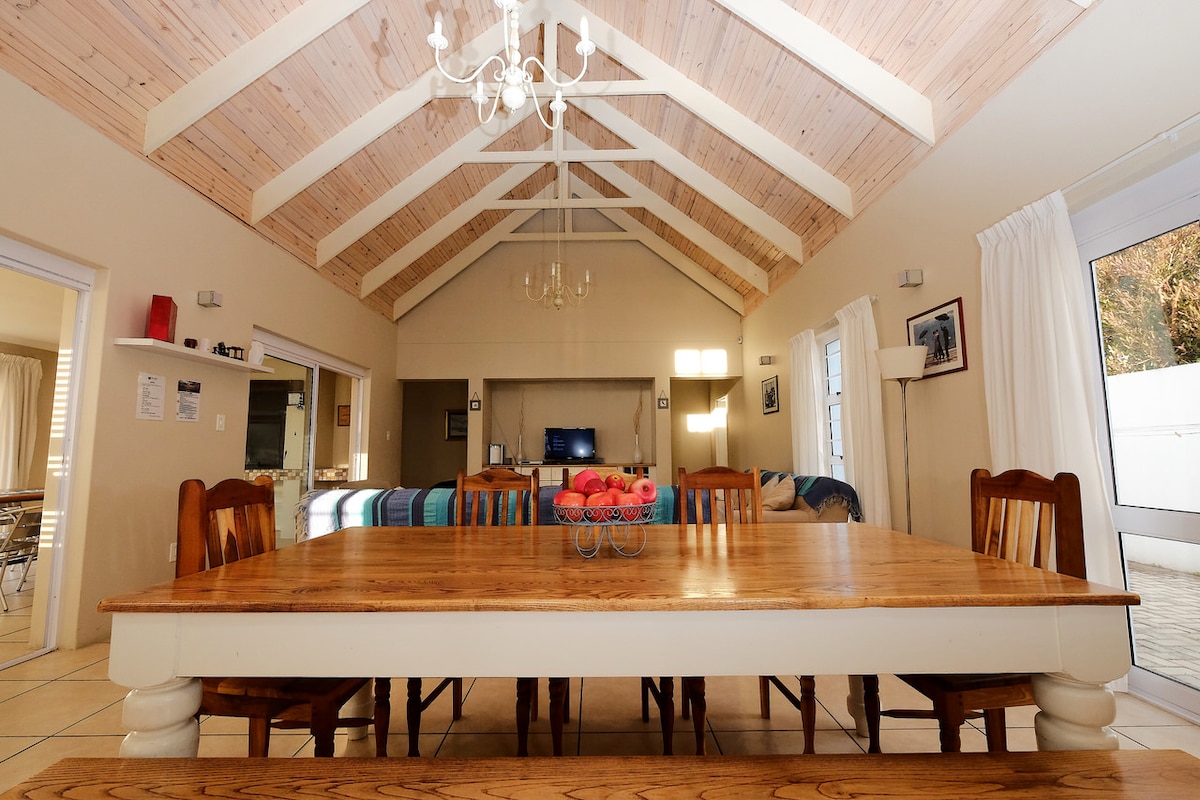 A spacious dining area is highlighted by a large wooden table surrounded by several wooden chairs. Exposed beams in the high ceiling enhance the room's open feel, while large windows allow natural light to fill the space. A chandelier hangs centrally above the table.