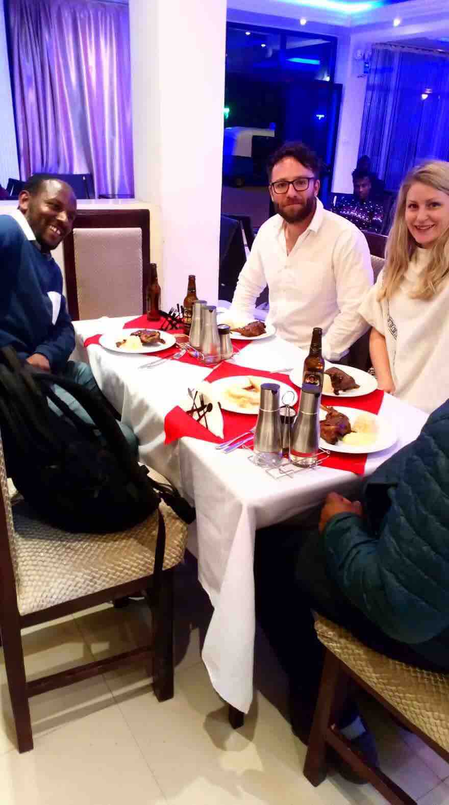 A dining table is set with white and red linens, showcasing plates of food and beverages. Four individuals are gathered around the table, with two still seated and engaged in conversation. The atmosphere is enhanced by soft lighting and elegant decor in the background.