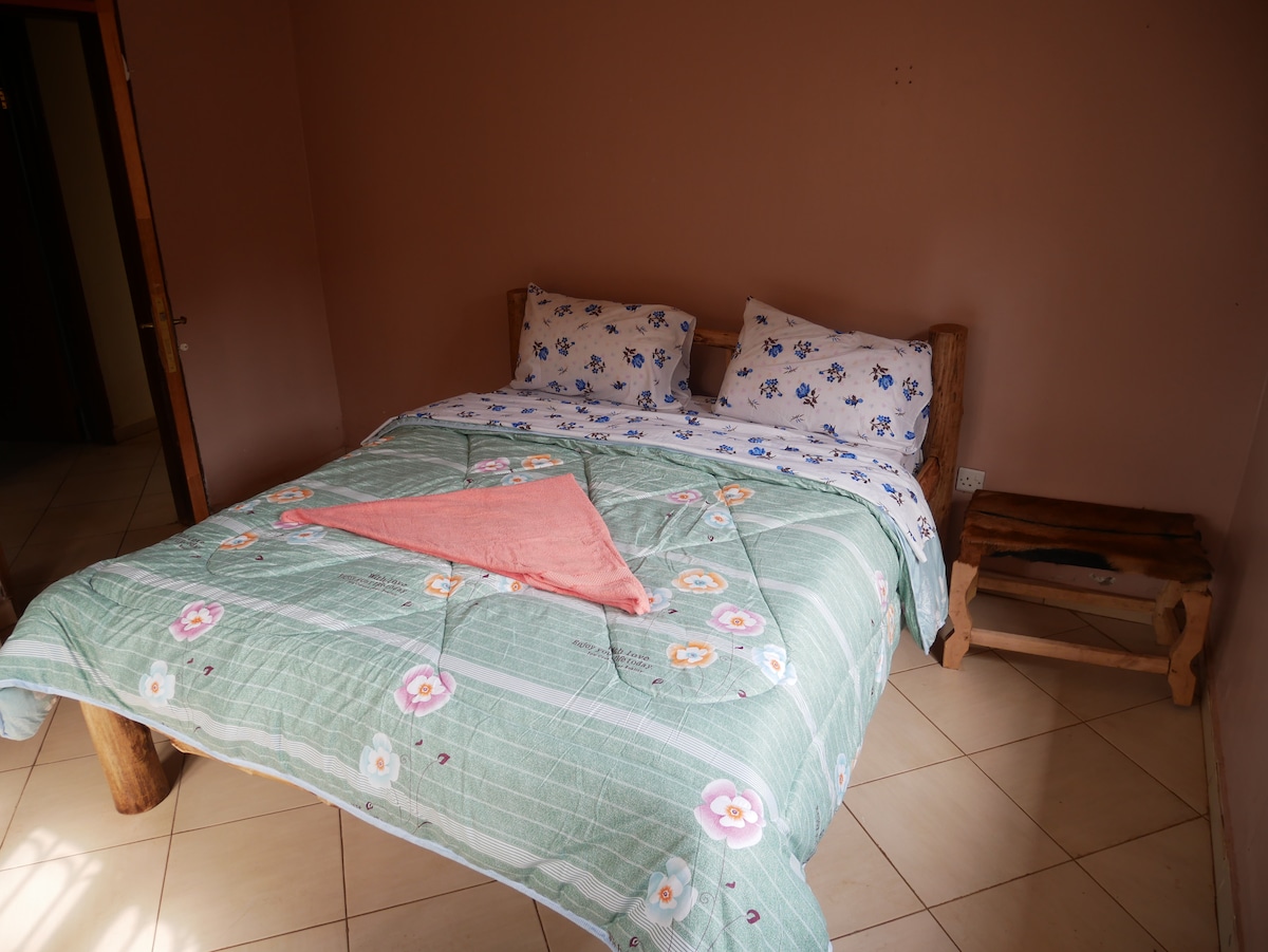 A spacious bed is presented with floral-patterned linens, complemented by decorative pillows. A wooden bedside table is visible beside the bed. The room features soft, neutral walls and tile flooring, creating a comfortable atmosphere.