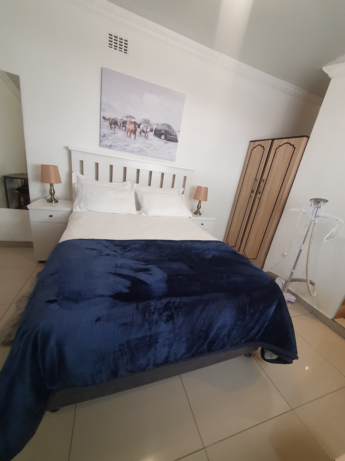 A cozy bedroom features a spacious bed with a rich navy blanket draped over it. Bedside tables with lamps are placed on either side, and a stylish wardrobe provides storage. A piece of artwork depicting a snowy landscape adorns the wall above the bed.