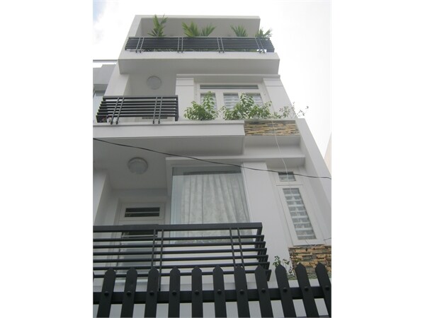 A three-story modern building is showcased, featuring multiple balconies adorned with potted plants. Large windows provide natural light, and sleek horizontal railings add a contemporary touch. A dark picket fence lines the front, creating a charming contrast with the white exterior.