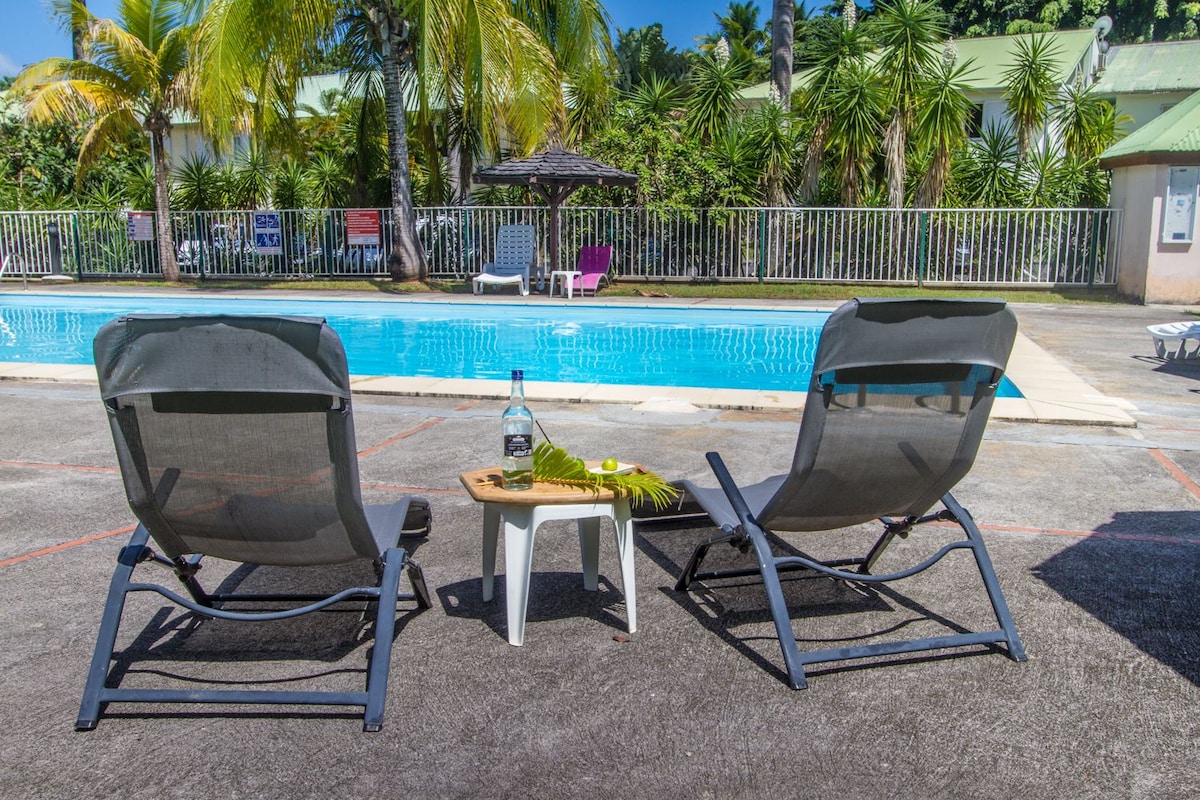 Two reclining lounge chairs are positioned beside a swimming pool, with a small table featuring a bottle and a slice of lime. Lush greenery and palm trees surround the area, providing a tranquil and relaxed setting.