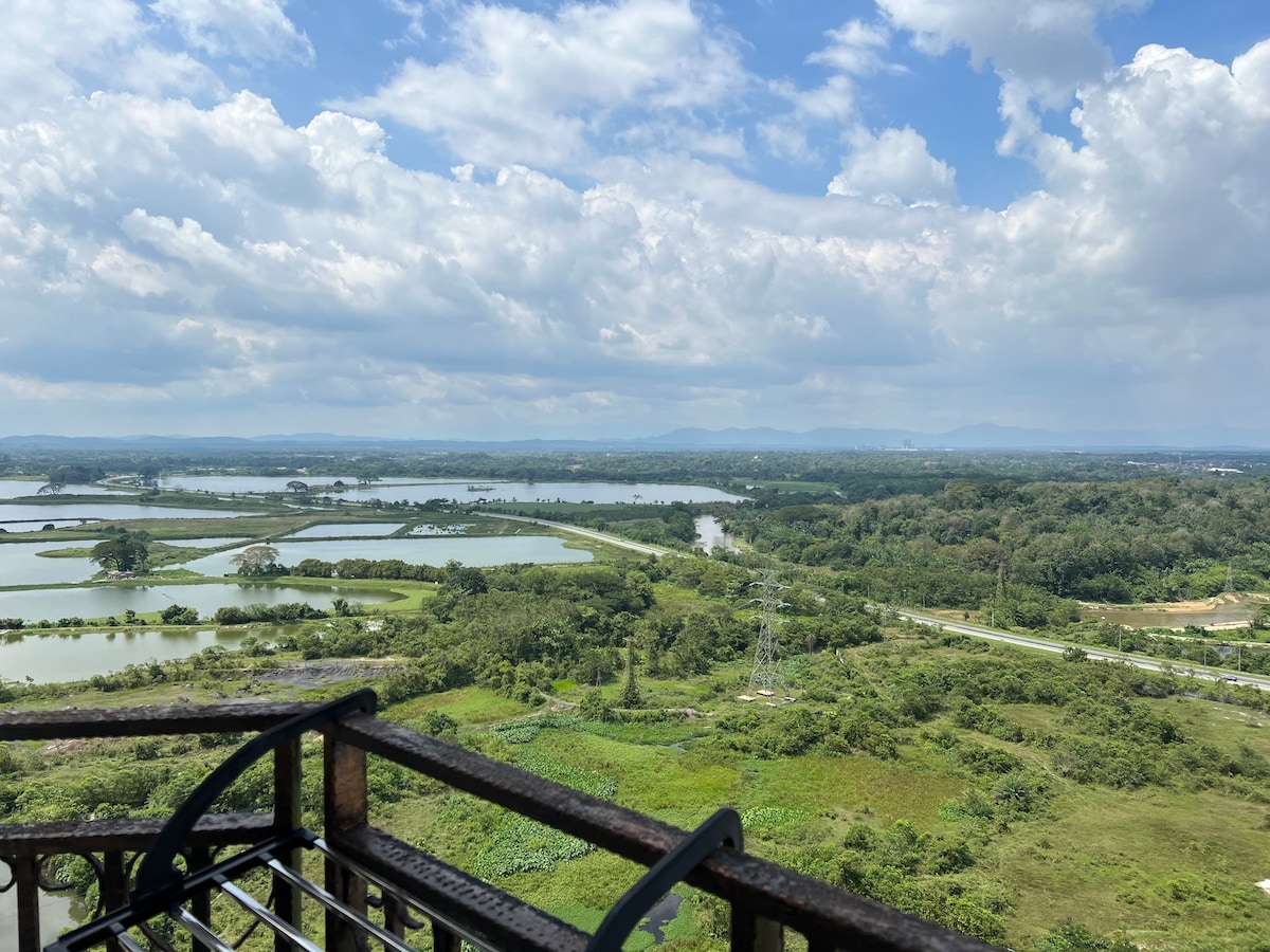 A panoramic view from a balcony showcases lush green fields and waterways stretching into the distance. The landscape is dotted with trees, and gentle hills are visible under a partly cloudy sky, creating a serene and natural setting.