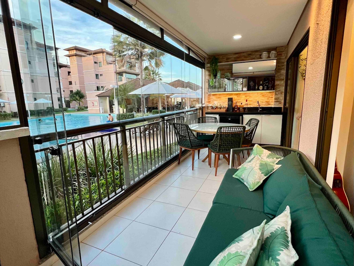 A glass-enclosed balcony features a seating area with green cushions and a round dining table. The view includes a swimming pool surrounded by greenery. Soft lighting enhances the inviting space, with wood accents visible on the walls behind the bar area.