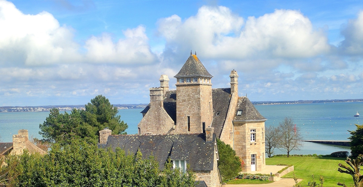 A charming stone building features distinctive architectural elements, including turrets and chimneys, set against a backdrop of serene blue waters. Lush greenery surrounds the structure, enhancing the picturesque setting and providing a glimpse of the coastal landscape.