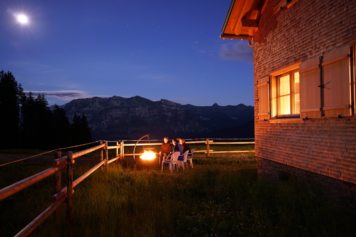 An intimate fire pit area is visible, surrounded by four white chairs. Soft light glows from the windows of the nearby chalet, while the moon and stars light up the sky above the distant mountains, enhancing the serene evening atmosphere.