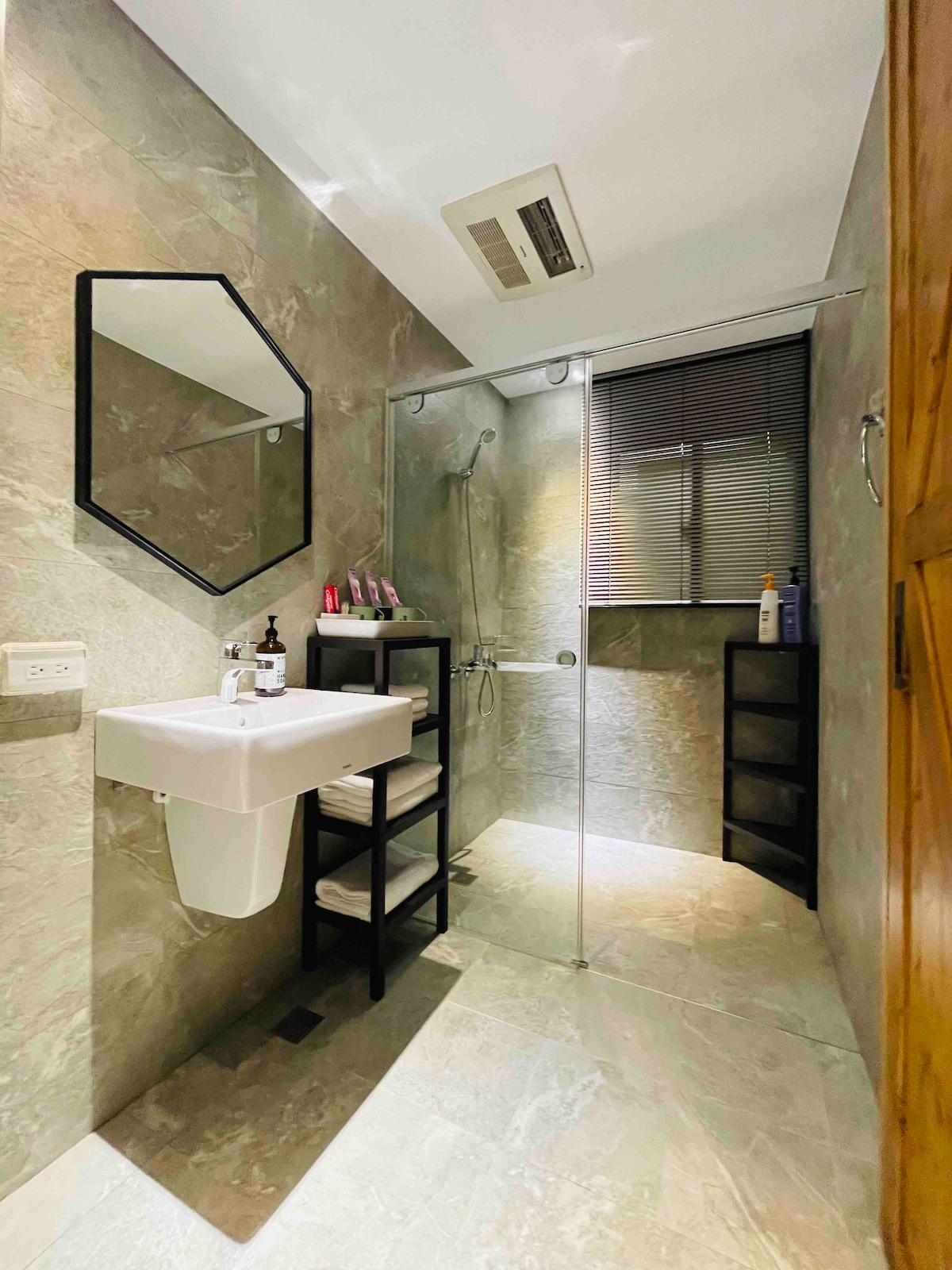 The bathroom features a spacious layout with natural stone-like tiling. A modern sink is positioned beside a sleek glass shower. An adjustable mirror adds functionality, while a black shelving unit provides storage for toiletries. Natural light enters through a window with blinds.