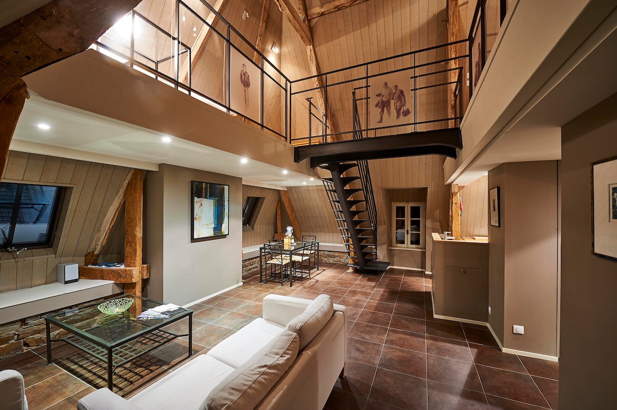 The open plan living area features a spacious seating arrangement with light-colored sofas and a glass coffee table. A dining area with metal chairs is visible, along with a staircase made of iron leading to the upper floor. Large windows provide ample natural light.