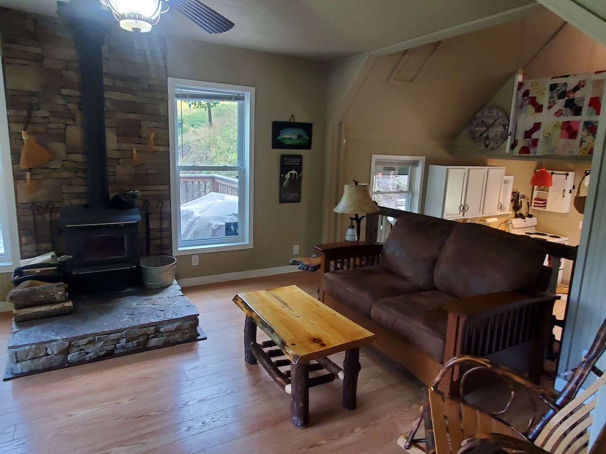 A rustic living area features a stone fireplace, wooden furniture, and large windows offering views of the surrounding greenery. A comfortable couch and a wooden coffee table create an inviting space, while art and decor add character to the walls.