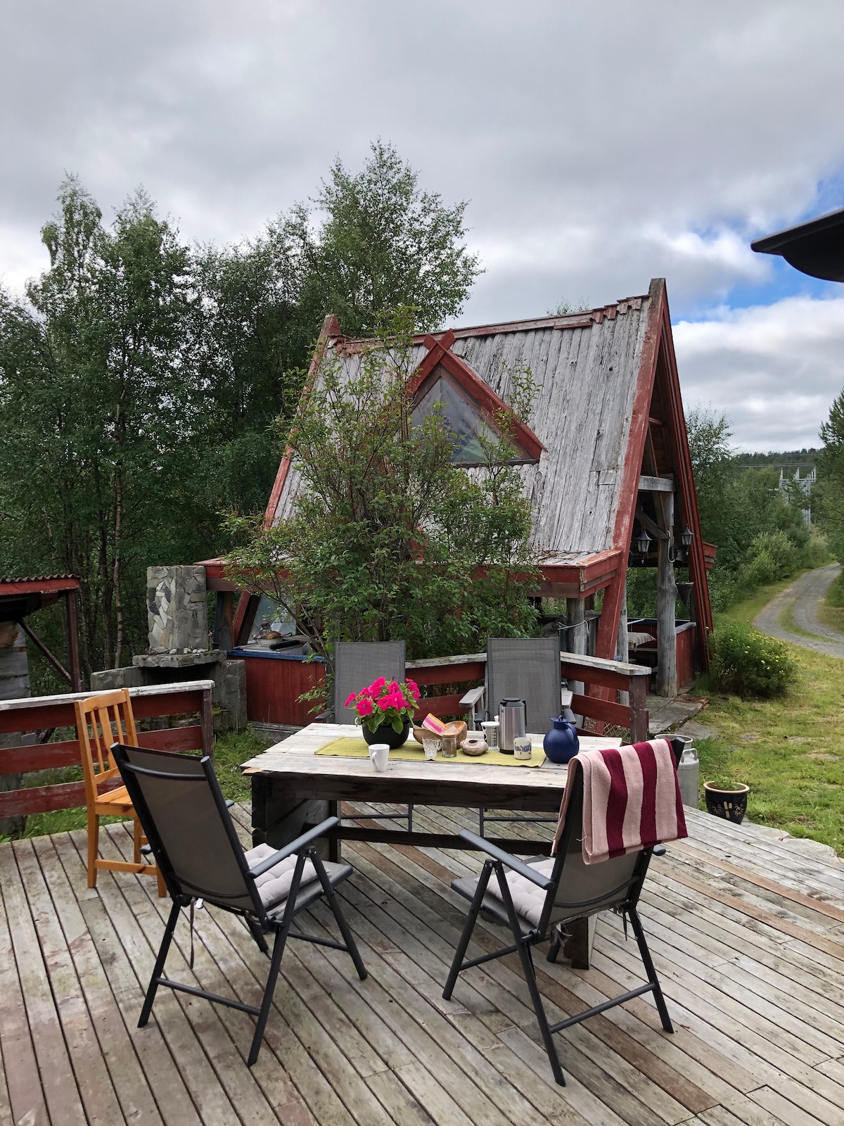 An outdoor dining area is displayed with a wooden table set for a meal, featuring floral arrangements and drinkware. Folding chairs surround the table, while a rustic A-frame structure with a weathered exterior is in the background, framed by greenery and a gravel path.